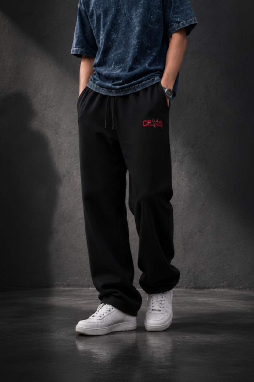 A person wearing black sweatpants with a small red logo on the left thigh, paired with a blue marled t-shirt and white sneakers, standing against a dark textured studio wall.